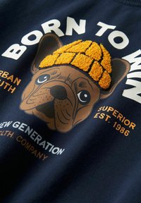 Bulldog francés con un gorro de punto naranja texturizado que lleva texto en blanco, naranja y azul sobre un fondo de tela oscura.