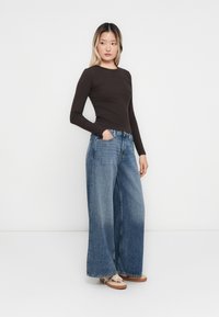 Ung kvinne med langt hår iført en tettsittende mørkebrun langermet topp, lysblå vide jeans og beige sko, står med hånden i lomma.