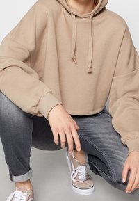 Sudadera beige recortada con cordones, mangas anchas, combinada con jeans grises y zapatillas rosas claras con cordones blancos y acentos negros.