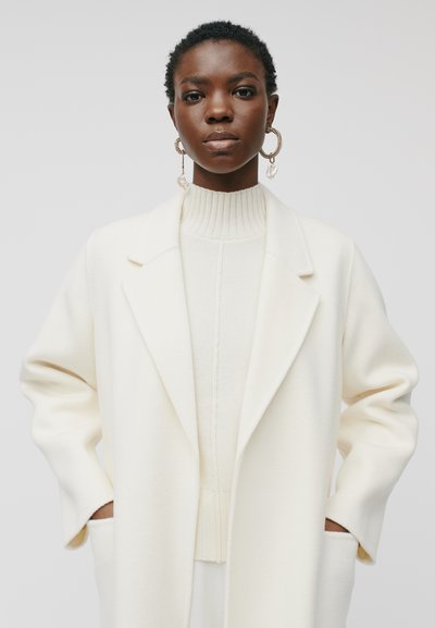 Weißer Wollmantel mit einem strukturierten Kragen und weiten Ärmeln, darüber eine helle, cremefarbene, gerippte Strickjacke. Markante Ohrringe vervollständigen den Look.