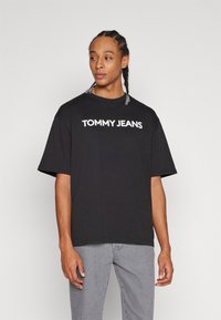 Camiseta de algodón negra con mangas cortas y cuello redondo, con el texto blanco "TOMMY JEANS" en el pecho. Llevada con jeans grises.