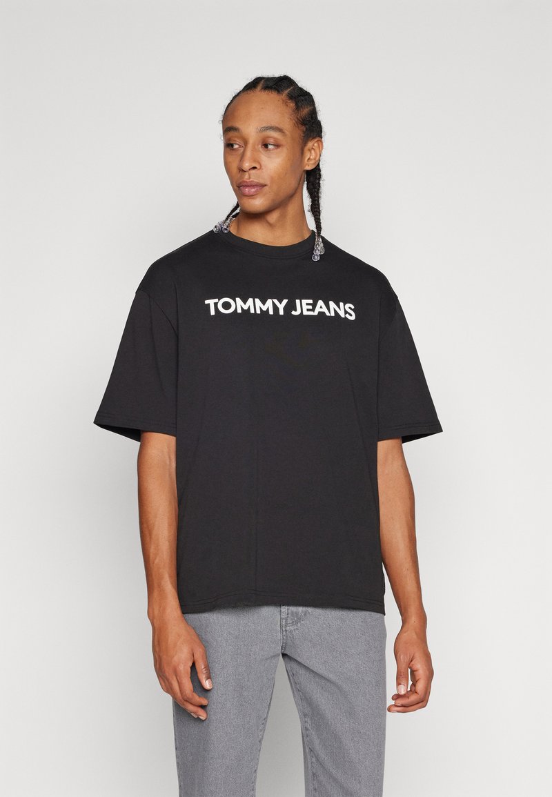 Camiseta de algodón negra con mangas cortas y cuello redondo, con el texto blanco "TOMMY JEANS" en el pecho. Llevada con jeans grises.