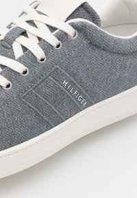Sneaker in tessuto grigio con lacci e suola bianchi, dotata di logo Hilfiger ricamato sul lato vicino ai fori metallici.