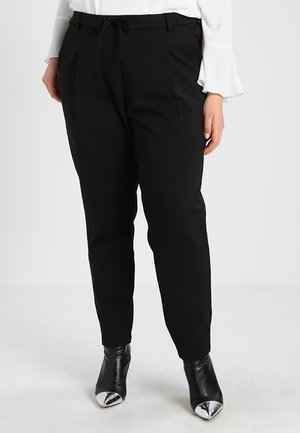 Femme portant un pantalon noir fuselé avec taille à cordon, blouse blanche avec poignets volantés, et bottines noires avec bouts pointus métalliques.