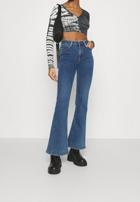 Blaue Schlagjeans mit hoher Taille, kombiniert mit einem verkürzten Oberteil aus Schwarz-Weiß-Tie-Dye mit langen Ärmeln. Das Model trägt klobige schwarze Stiefel.