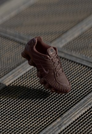 Dunkelbrauner Sportschuh mit strukturierter Sohle und Schnürdesign, der auf einer gemusterten Metalloberfläche ruht.