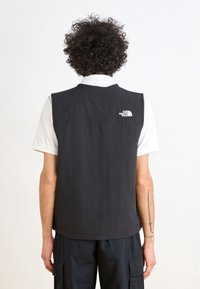 Chaleco negro sin mangas hecho de tela con textura suave, con un logo blanco en la parte superior de la espalda, usado sobre una camisa con cuello.