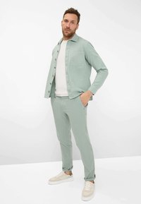 Hellgrüne, knöpfbare Bluse mit einer Tasche, kombiniert mit passenden schmal geschnittenen Hosen. Weißes T-Shirt mit Rundhalsausschnitt darunter. Beigefarbene Sneakers.