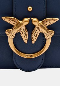 Borsa a mano in pelle blu scuro con chiusura circolare in tonalità oro, decorata con due uccelli stilizzati, dettagliate motivi di piume e un'iscrizione del logo.