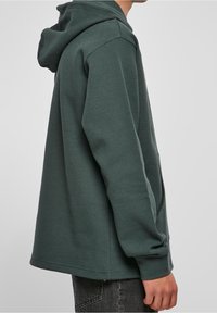 Felpa verde con cappuccio realizzata in materiale a maglia testurizzata. Presenta un cappuccio, maniche lunghe e una vestibilità rilassata. Nessuna grafica o decorazione visibile.