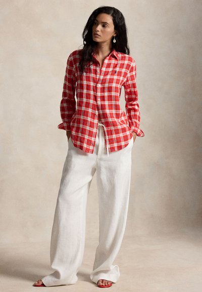 Kvinne som står iført en rød- og hvitternet langermet skjorte, vide hvite bukser og røde sandaler mot en ensfarget beige bakgrunn.