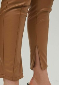Pantalons en simili cuir marron avec une coupe ajustée, dotés de détails de couture verticaux et d'une petite fente à l'ourlet. Texture lisse et brillante.