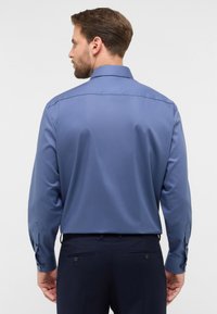 Camicia blu a maniche lunghe con colletto classico, texture liscia e polsini con bottone; vista da dietro, che mette in mostra la vestibilità su misura e il giogo posteriore.