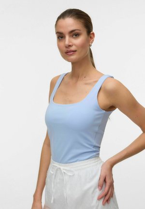 Woman with light brown hair in a ponytail wears a light blue sleeveless top and white drawstring pants, posing with one hand on her hip.