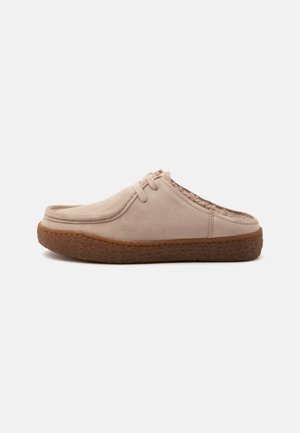 Beige suede slip-on shoe with brown textured sole, round toe, closed front, open heel, and minimal laces, shown on a plain background.
