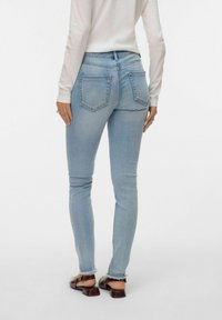 Šviesiai mėlynos siauro kirpimo džinsai su aukštu liemeniu, dviem kišenėmis gale ir nusitrynusiais kraštais, derinami su ruda avalyne ant vienspalvio fono.