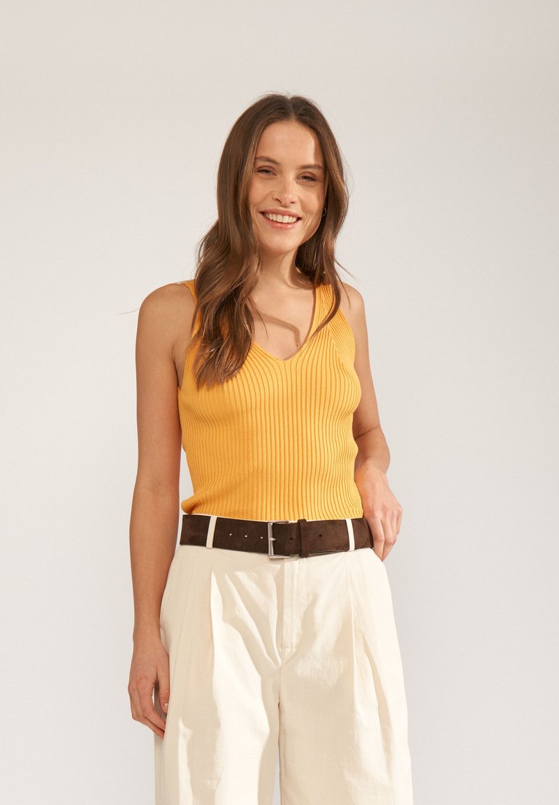 Femme souriante aux longs cheveux bruns portant un top jaune sans manches à côtes, un pantalon blanc plissé et une ceinture marron foncé, debout devant un fond uni.