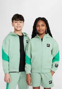 Gröna zip-up hoodies med svarta Nike Air-loggor, med mörkare gröna accenter. Båda bär matchande shorts med liknande loggor och färger.