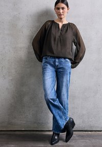 Schwarze transparente Bluse mit einem Reißverschluss vorne, kombiniert mit hellblauen Schlagjeans und schwarzen Stiefeletten, vor einem Betons Hintergrund.