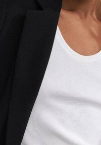 Mustapukuiset bleiseri valkoisen ribatun topin päällä, jossa on pyöreä kaula-aukko. Kankaat ovat sileäpintaisia, ja niissä on näkyviä hienovaraisia yksityiskohtia.