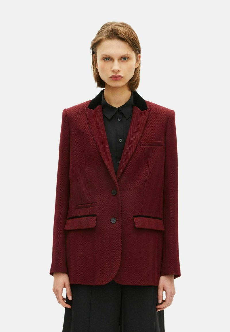 Blazer bordeaux con colletto a rever, dettagli in velluto nero, due tasche frontali, chiusura a un bottone e tessuto testurizzato. Vestibilità su misura.