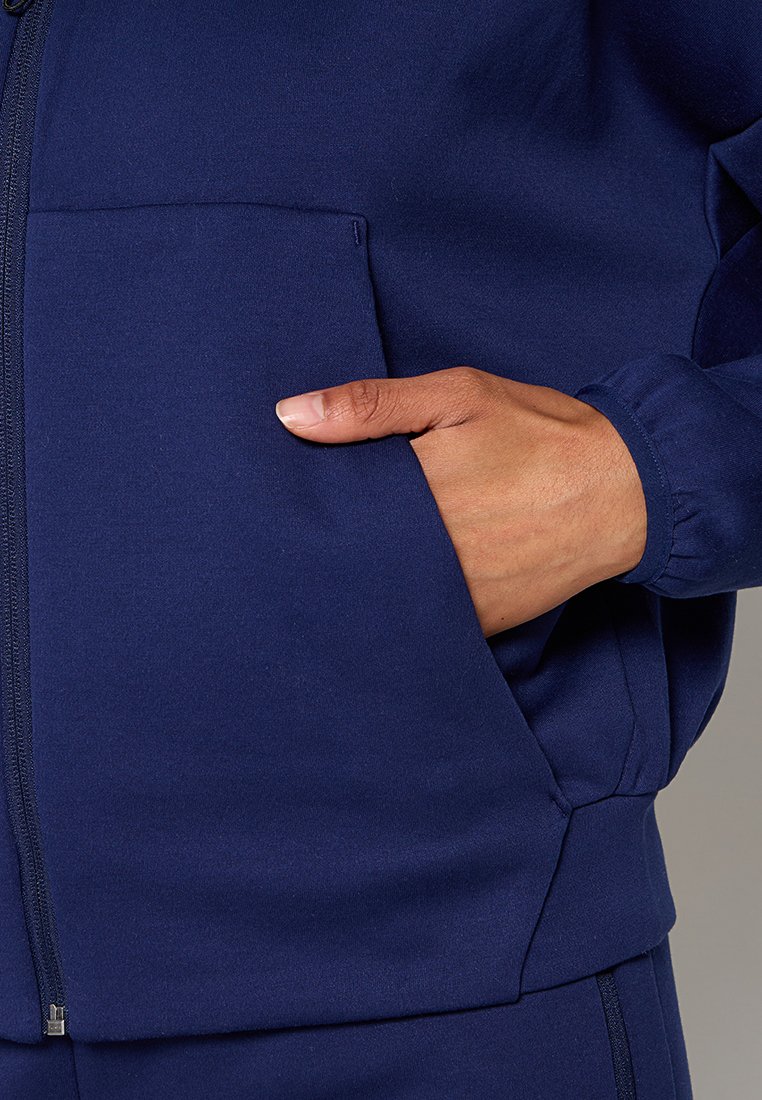 Navyblaue Zip-Jacke aus glattem Stoff, mit einer Vordertasche und elastischen Bündchen, sichtbaren Nähten entlang des Reißverschlusses.