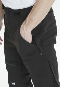 Svarta byxor tillverkade av stretchmaterial har en dragkedjeförsedd sidficka och en platt midja med knapp- och kardborrefästen, strukturerad yta.