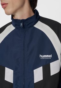 Marindrabbad sportjacka i marinblått med vita och svarta diagonala detaljer, dragkedjestängning och "hummel sportswear"-logotyp på bröstet. Lättviktigt tyg.