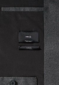 Tissu de veste gris foncé à texture lisse. Comprend des étiquettes noires marquées "MNG" et "SLIM FIT" cousues sur la doublure.