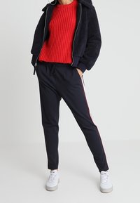 Veste polaire bleu marine à fermeture éclair avec capuche, portée sur un pull rouge en maille texturée. Pantalon noir avec des bandes blanches et rouges sur le côté. Baskets blanches à semelles épaisses.