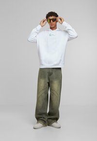 Sudadera blanca con un bolsillo frontal y un logo verde, combinada con unos vaqueros de corte amplio en verde claro que presentan detalles de costura curvada y zapatillas blancas.