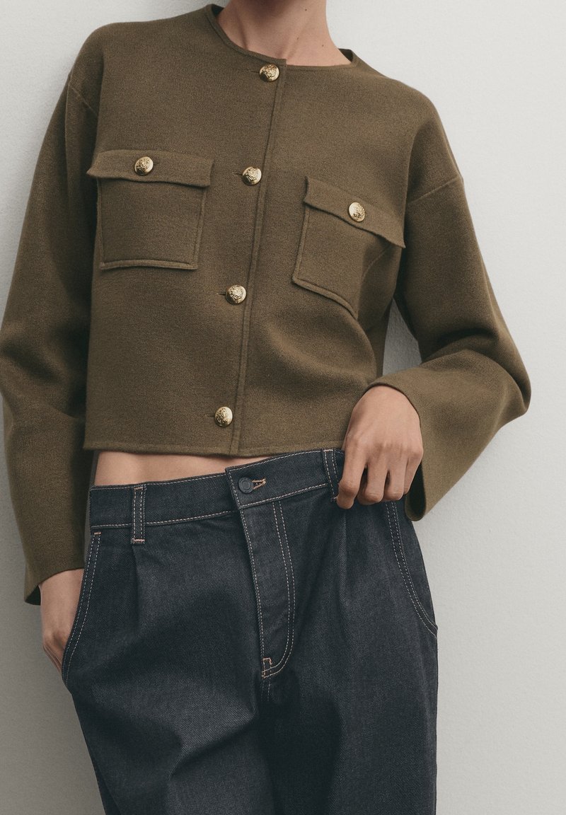 Olive green cropped sweater with gold buttons and two front pockets, paired with dark blue denim high-waisted trousers featuring contrasting stitching.