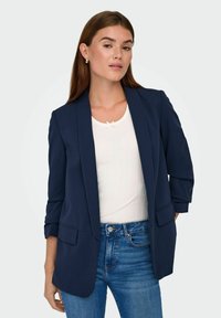 Kvinde iført en marineblå blazer over hvid top og blå jeans, stående med den ene hånd bag ryggen mod en ensfarvet baggrund.