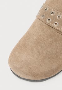 Tan suede boot with a rounded toe, featuring a strap with circular metal grommets. Soft texture and black rubber sole.