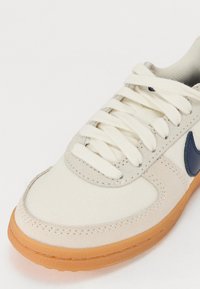 Gros plan d'une basket beige et blanche avec des lacets blancs, une semelle en gomme et un logo bleu foncé sur le côté.