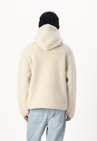 Felpa in pile color crema con una superficie morbida e текстurizzata, dotata di un ampio cappuccio e di una vestibilità comoda. Indossata con jeans blu chiaro.