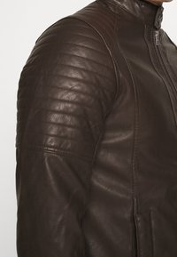 Giacca in pelle marrone con dettagli trapuntati sulle spalle, colletto alto e zip laterale. La texture è liscia con una leggera lucentezza.