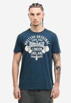 Mann mit geflochtenem Haar und Arm-Tattoos trägt ein blaues Lonsdale London England T-Shirt mit Boxhandschuhe-Logo und steht vor einem weißen Hintergrund.