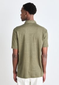 Kortärmad polo i olivgrön färg, med en strukturerad tyg, krage och rak nederkant, sedd bakifrån.
