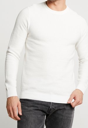 Weißer Strickpullover mit gerippter Textur, rundem Ausschnitt und langen Ärmeln, mit einem figurbetonten Saum. Kombiniert mit dunkelgrauen Jeans.