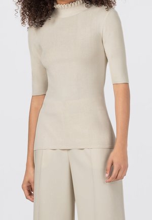 Vrouw in een beige top met korte mouwen en een geplooide hals, met bijpassende beige wijde broek, staand tegen een effen achtergrond.