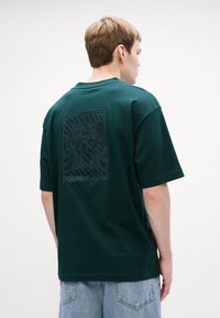 Mørkegrønn t-skjorte med kort erme, med et teksturert grafisk design på baksiden og en avslappet passform. Laget av bomull med en jevn tekstur.