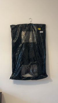 Sacca per abiti nera appesa a una gruccia metallica contro un muro beige, con etichette di spedizione visibili sulla sacchetta.