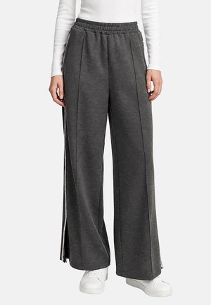 Pantalones anchos gris oscuro con cremalleras laterales, combinados con deportivas blancas y una camiseta blanca de manga larga, mostrados de la cintura a los pies.