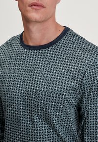 Marineblaue Langarmbluse mit geometrischem Muster, Rundhalsausschnitt und Brusttasche. Der Stoff ist weich mit einer strukturierten Oberflächenbeschaffenheit.