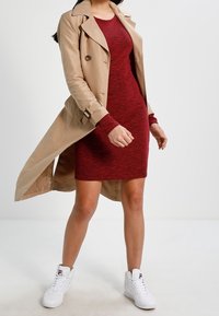 Un trench-coat beige sur une robe rouge à côtes, avec de longues manches et une coupe ajustée. Porté avec des baskets montantes blanches.