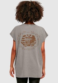 Grå oversized t-shirt lavet af blødt stof, prydet med et stort brunt ægyptisk inspireret design med hieroglyffer på ryggen.