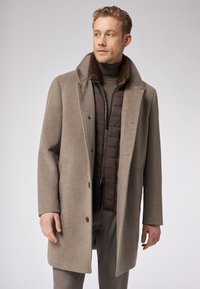 Cappotto in lana marrone con un ampio colletto, abbinato a uno strato interno trapuntato marrone scuro. Presenta una chiusura con bottoni e una texture liscia.