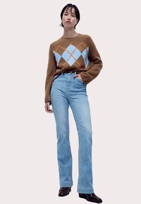 Pull en maille marron avec motif en losanges bleu et orange, associé à un jean évasé bleu clair. Bottines noires. Fond neutre.
