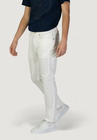 Jeans bianchi slim-fit con dettagli in patch, multiple tasche e accessori in tonalità oro, abbinati a sneakers bianche e una maglietta grafica blu navy.